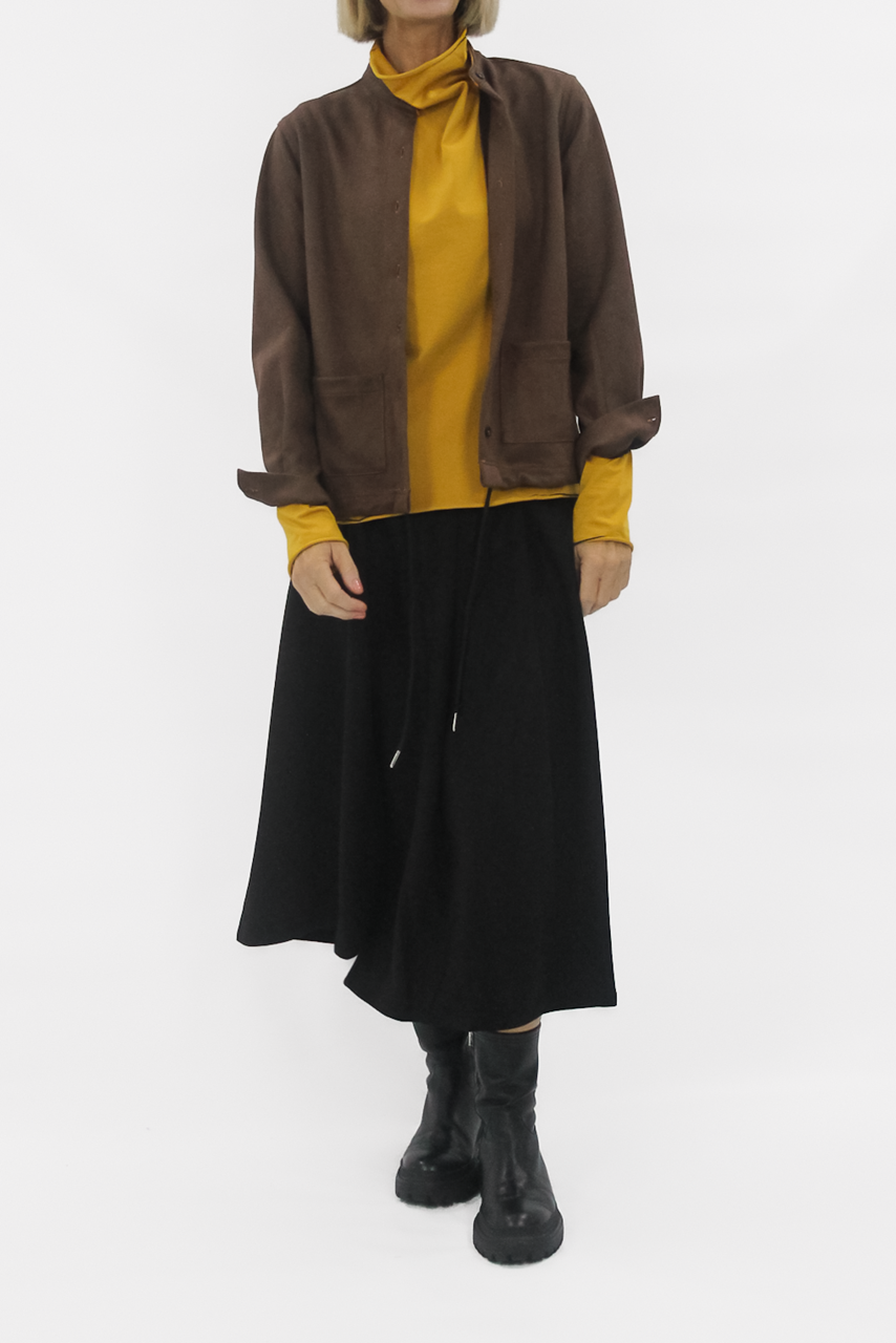 Person wearing a brown jacket, mustard shirt, black skirt, and black boots on a white background