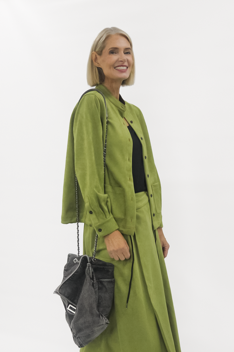 Woman wearing a green vegan suede jacket holding a black handbag against a white background